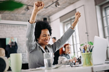 Cheerful African American businesswoman celebrating success while using computer in the office. Low angle view of happy black businesswoman working in the office and celebrating her success with raised arms.